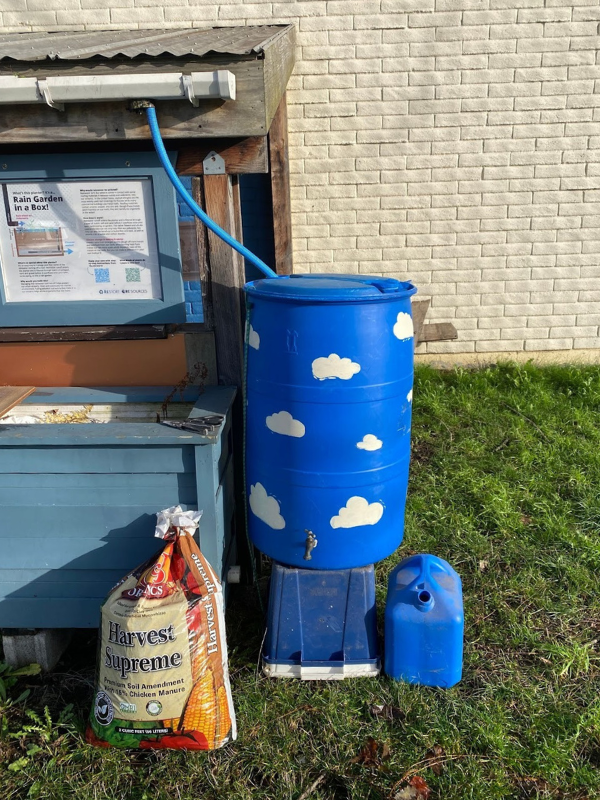 finished project of DIY rain barrel set up next to a bike shed at The RE Store. With a bag of soil leaning up against a garden box as a display